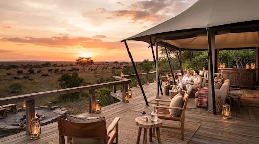 Bespoke luxury tented safari lodge lounge deck overlooking the Serengeti acacia plains at sunset, stylish canvas chairs, ambient brass lanterns glowing, teak wood deck, African wildlife silhouette in distance, luxury safari atmosphere.