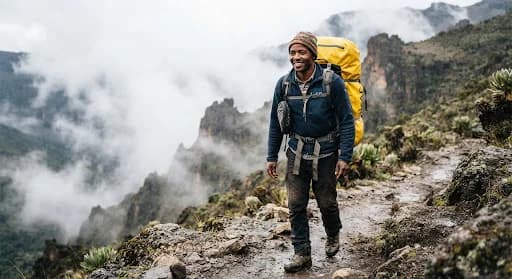 Heartwarming authentic documentary photograph of a smiling mountain porter on Mount Kilimanjaro wearing warm high-performance fleece and waterproof duffel backpack, mist rising behind him on the Machame ridge, respectful human-centric framing.